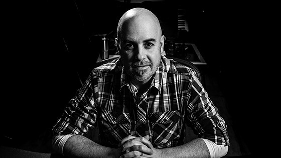 Grad Terry Watson sits with his hands clasped at a desk. He is wearing a plaid button-down shirt and is in a music studio.