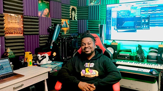 Grad Shawn Deshield smiling while seated at a desk in a home studio featuring a keyboard, audio equipment, and a large screen displaying audio software while wearing a black hooded sweatshirt with a colorful design.