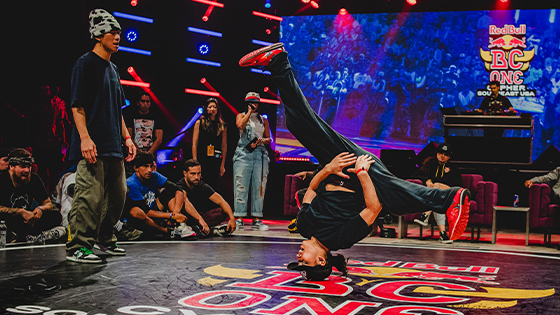 A dancer is doing a hands-free headstand with their legs spread while their competitor, the audience, and the judges watch.