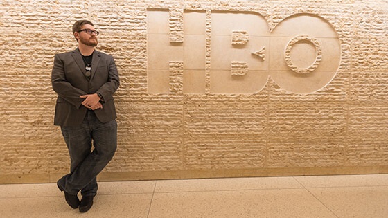 Wearing a suit and glasses, Stephen Beres leans against a beige, textured stone wall with the HBO logo carved into it.