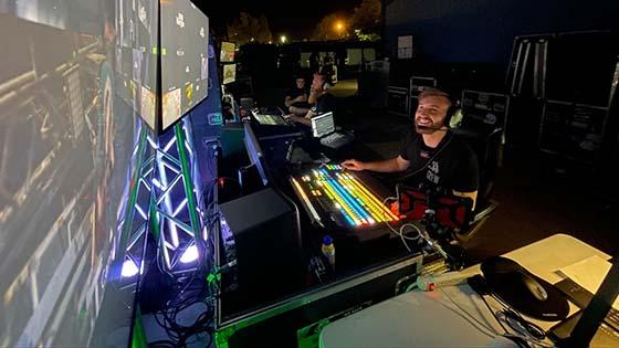 A smiling young man wearing headphones and sitting at a video switcher looks up at two large video screens in a production studio.