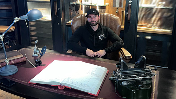 Rob Prager sits in a leather chair behind a large desk. He is smiling and wearing a black baseball cap and a black hoodie.