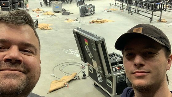 Two smiling men with beards stand in a backstage area. There are wires on the floor behind them, as well as an open trunk filled with production equipment.