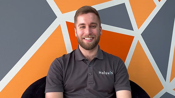 A man with light hair and a beard wearing a grey Holovis polo while seated and smiling in front of an orange, grey, and white geometric background.