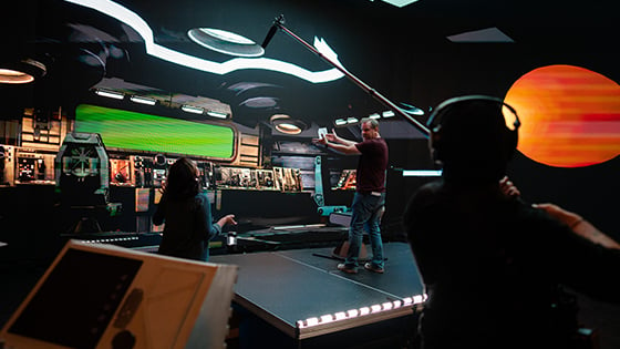 Space Pups’ director Jason Murphy stands onstage in a virtual spaceship set. He is talking to a crew member while another crew member holds a boom mic.