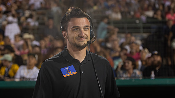 Full Sail grad Joe Zollo wears a headset on the field as Show Coordinator for the Savannah Bananas, with fans filling the stands behind him.