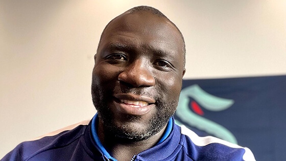 Grad Lamont Buford sports a blue jacket with a Seattle Kraken logo while smiling and holding a camera.