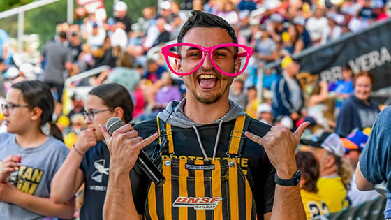 A man standing and looking excited while holding a microphone in oversized pink glasses and yellow and black striped overalls in the foreground of a large crowd at a stadium.