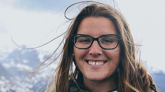 Full Sail grad SaJorden Miller pictured from the shoulders up, in the background is a snowcapped mountain range. She is smiling, wearing a green zip-up jacket with orange accents and black framed glasses.