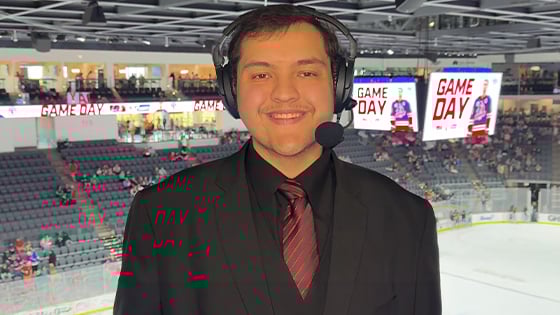 A smiling sports broadcaster in a suit and headset stands in an arena booth above an ice hockey rink with "Game Day" on screens behind.