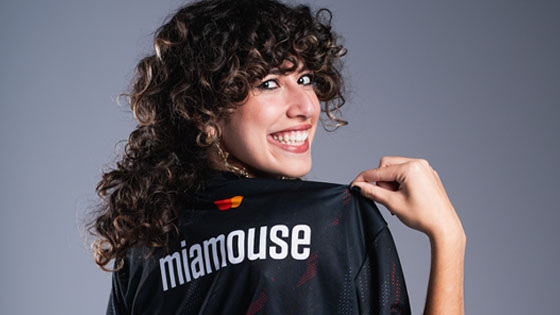 A smiling woman with curly hair holds up a jersey that reads “miamouse” on the back, showing it proudly to the camera.
