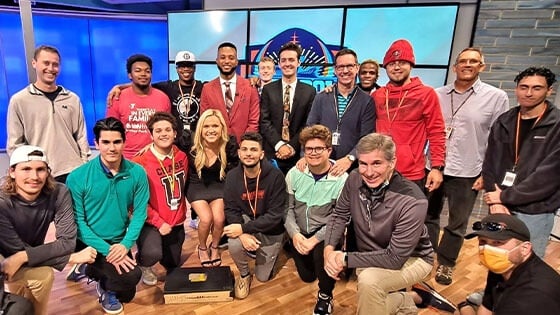 A group shot of several Full Sail students and instructors in front of a large screen on set at the Dan Patrick School of Sportscasting.