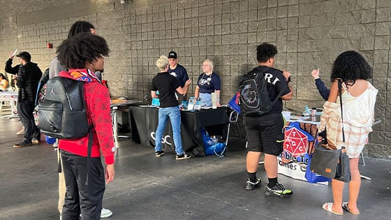 Several students gather on a patio with various tables set up. Club representatives stand at the tables talking with students.