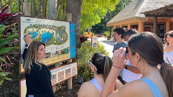 Crystal O’Hea stands at a map of Discovery Cove and speaks to students as they stand around.