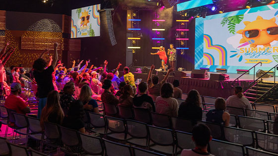 Several students raise their hands attentively at the Summer Sizzle as two staff members call on participants. The large room is colorfully lit up and looks very lively.