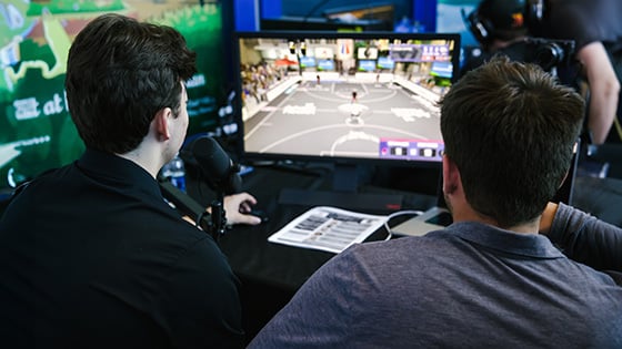 Two people have their backs to the camera while watching ‘NBA 2K’ gameplay on a small monitor on a table in front of them.