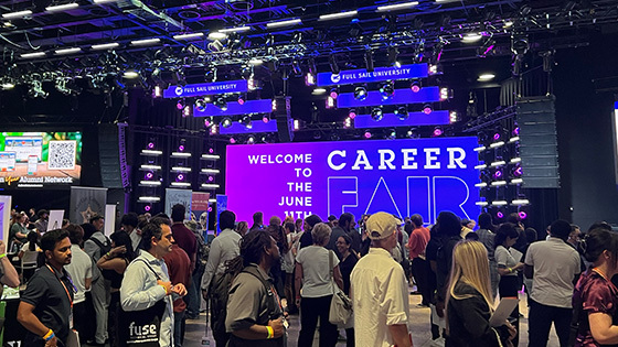 Students and alumni stand around and talk to potential employers. A screen behind them displays, “Welcome to the June 11th Career Fair.”