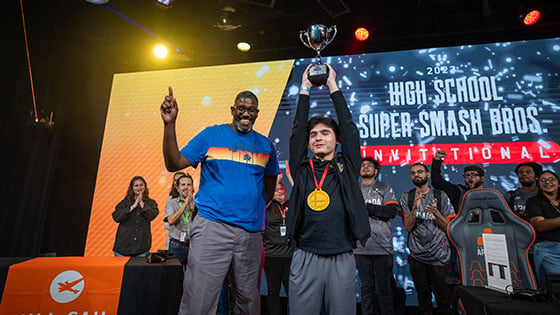 Two people are standing side by side on a stage while one holds a large silver trophy over their head.