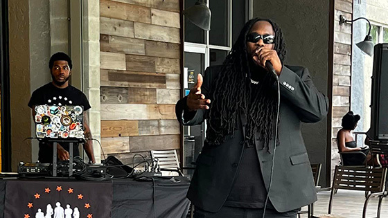 Guest speaker Souljah Bless, dressed in an all black outfit, speaks into a microphone on a patio stage. Behind him, a DJ stands at a booth that reads “Student Community Association.