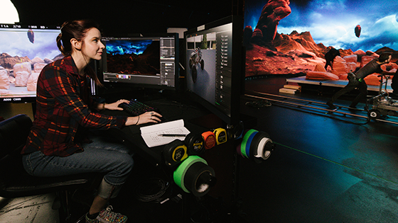 A woman is sitting at a desk in front of several monitors. She is in a virtual production studio that has been designed to look similar to Mars.