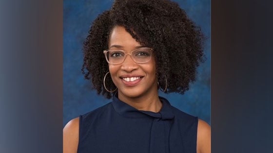 Tiana, an African American woman, sits smiling at the camera. She is wearing a sleeveless navy blue blouse, silver hoop earrings, and glasses with translucent beige frames.