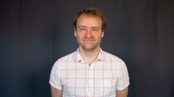 Paul Ellison smiles at the camera. He has short blonde hair and blue eyes and is wearing a white and red button-up shirt.