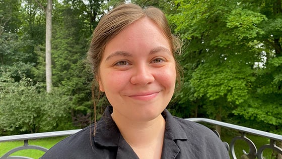 Emmi Litsenberger sits on a porch in front of a grassy lawn and trees. She is smiling and wearing a gray button-up shirt.