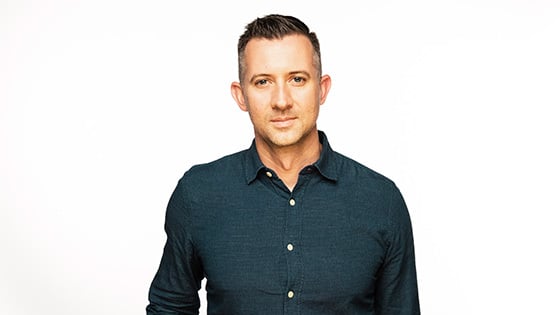 Dean Sell stands in front of a white background. He is wearing a dark button down shirt.