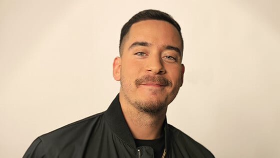 Eric Medina smiles at the camera. He is wearing a black zip-up jacket, black t-shirt, and gold chain necklace.