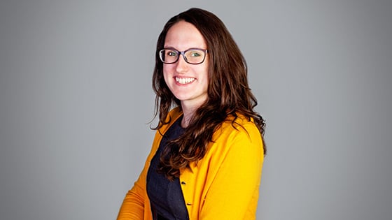 Tasha Rosenbaum smiles at the camera. She has brown hair and is wearing glasses, a yellow cardigan, and a blue sweater.