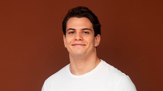 Gabriel Garnier smiles at the camera. He is wearing a white t-shirt that says “U READY?” and standing against a brown backdrop.