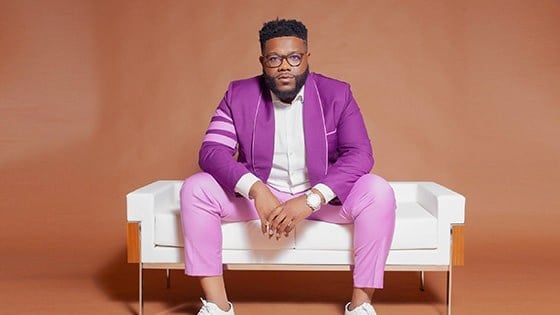 A man with black hair and a beard wearing glasses, a purple suit, and a white button down shirt while seated on a white couch against a brown background.