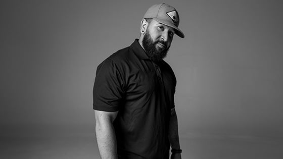 Kevin Hall stands and looks at the camera. He has a beard and is wearing a baseball cap, a collared short-sleeve shirt, and jeans.