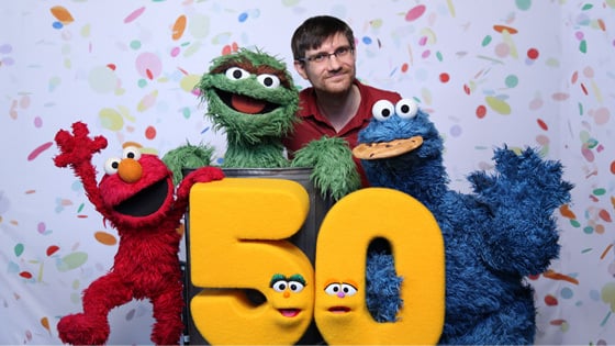 Chris Sassano stands with Oscar the Grouch, Cookie Monster, Elmo, and a puppet shaped like the number 50 with confetti behind them.