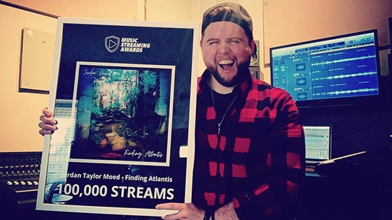 Jordan Moed stands in his studio and holds an award for 10,000 streams for his song “Finding Atlantis.”