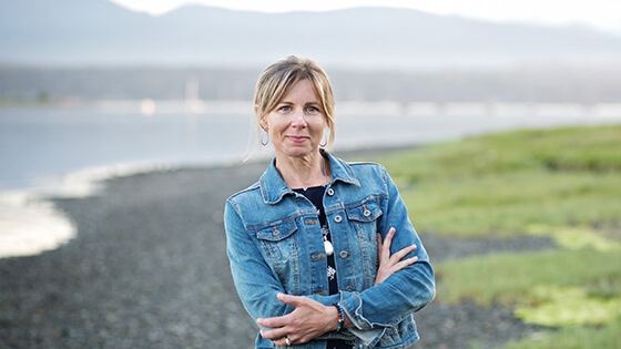 A blonde woman wearing a denim jacket over a navy top and black skirt stands with her arms crossed. She is outdoors in front of a river with mountains far in the background.