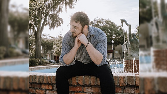 A man with short, dark, curly hair in a blue button down shirt and wireframe glasses looking to the side while sitting in front of a fountain.