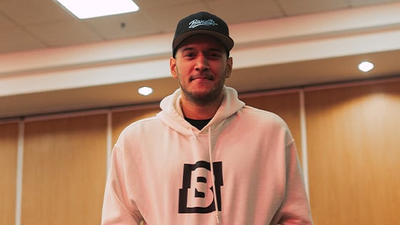 A man standing against a wood paneled wall in a white hooded sweatshirt featuring the Bandits logo and a black Bandits Gaming snapback cap.