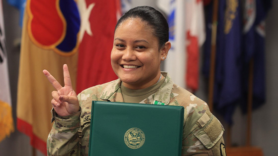 Anjelica Asejo smiles at the camera holding a certificate. She is wearing her Army uniform with a row of flags behind her as she holds her fingers in a peace sign.