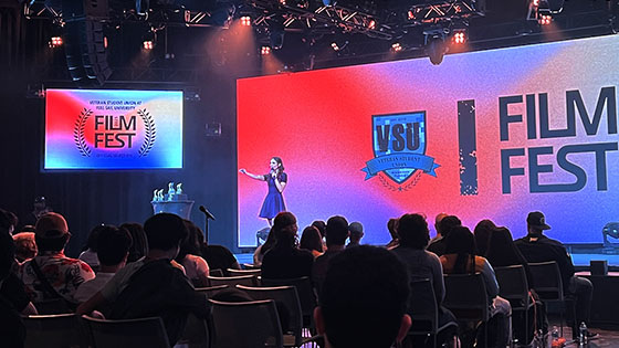 An audience faces a stage with a speaker holding a microphone. A large screen displays “Film Fest” and the VSU logo on a colorful background.