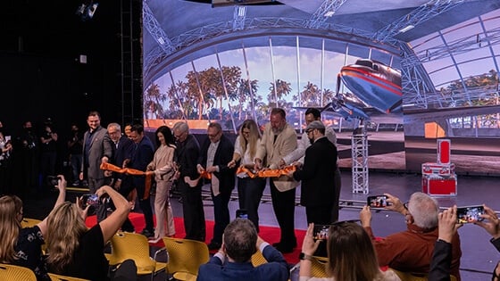 A group of eight men and two women stand on a red carpet in front of a large screen displaying an airplane and palm trees. They are each holding a pair of scissors and cutting a long orange ribbon. The seven audience members in front of them are photographing them with cell phones.