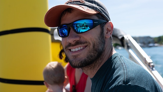 A man wearing a blue t-shirt, a blue and red baseball cap, and sunglasses with polarized lenses sits on a boat on a sunny day. He is smiling at the camera and there is a brunette woman and a blonde child sitting out of focus behind him.