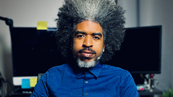 Bill Hudson sits in an office chair in front of a desk with two computer monitors. He has black and gray hair and wears a blue button-up shirt.
