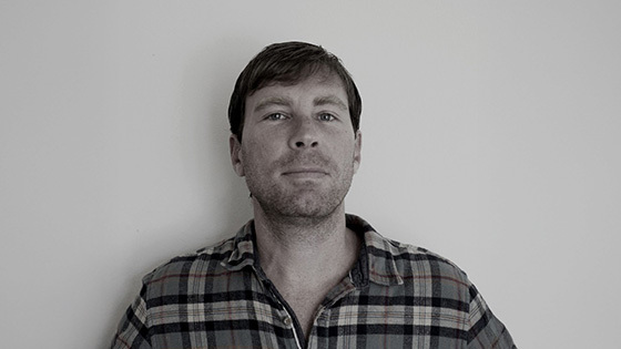 A man in a plaid shirt stands against a plain wall, looking directly at the camera with a very slight smile.