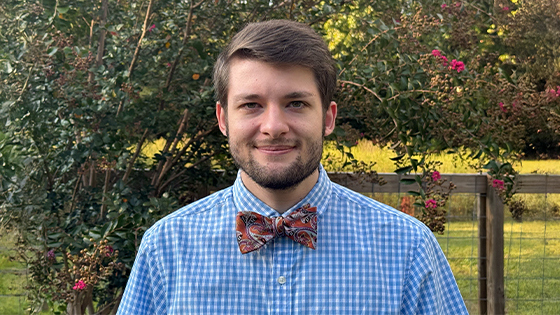 Noah White smiles at the camera in front of a row of green bushes with pink flowers. He is wearing a blue shirt and red bowtie.