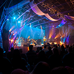 A band plays music in a large, dark auditorium with several bright blue and purple lights pointing at the stage.