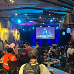 A crowd of students sit at tables in a large room. A large screen displays a video game and decorative paper lanterns hang around the room.
