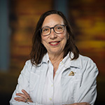 Abby Ginsburg smiles at the camera in front of a wood background. She is wearing a white jacket and glasses.