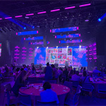 Groups of people sit around circular tables in a large room with pink and blue lights. A large screen displays photos and the American Cancer Society Making Strides Against Breast Cancer logo