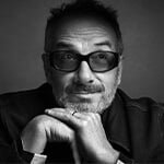 A black and white portrait of singer, songwriter, and producer Elvis Costello wearing dark-tinted glasses and a wedding band.